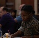 A U.S. Marine in woodland MARPAT camouflage uniform is seated at a table, focused on writing or filling out paperwork. The setting appears to be an indoor venue, possibly a library or community hall, with several civilians visible in the blurred background. The Marine is wearing gold-framed glasses and a smartwatch, suggesting a formal or administrative event.