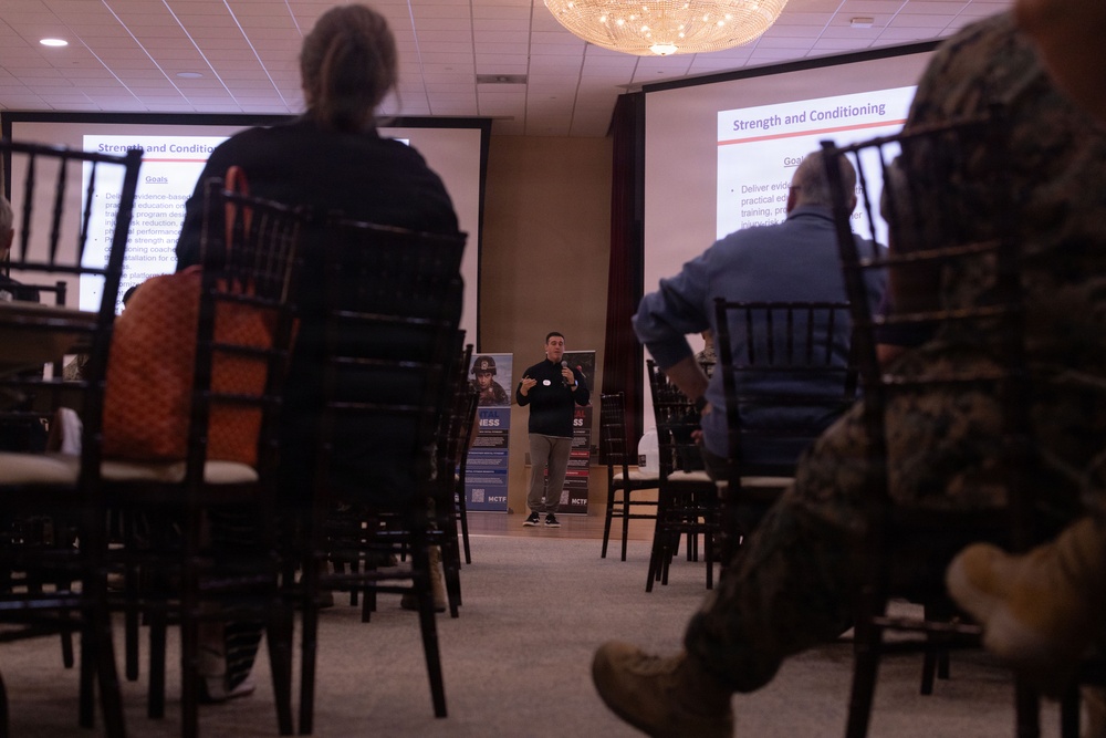 A presenter is delivering a 'Strength and Conditioning' presentation in a conference or banquet room setting. Two projection screens display the same slide titled 'Strength and Conditioning' with visible bullet points under a 'Goals' heading. The audience includes individuals in civilian clothing and at least one person in military camouflage uniform.