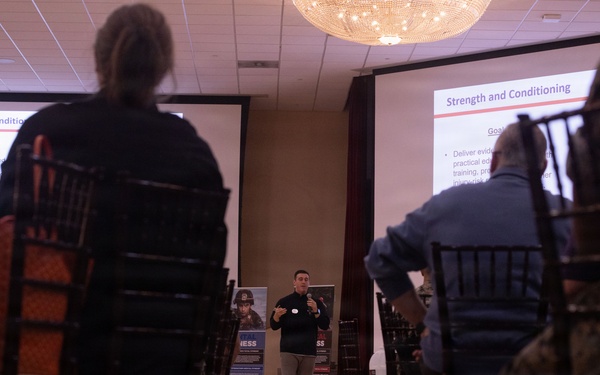 A presenter is delivering a 'Strength and Conditioning' presentation in a conference or banquet room setting. Two projection screens display the same slide titled 'Strength and Conditioning' with visible bullet points under a 'Goals' heading. The audience includes individuals in civilian clothing and at least one person in military camouflage uniform.