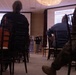 A presenter is delivering a 'Strength and Conditioning' presentation in a conference or banquet room setting. Two projection screens display the same slide titled 'Strength and Conditioning' with visible bullet points under a 'Goals' heading. The audience includes individuals in civilian clothing and at least one person in military camouflage uniform.