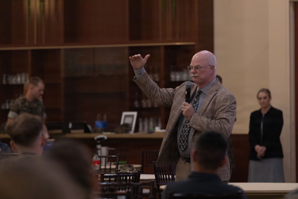 A middle-aged man in a plaid blazer and patterned tie is speaking to an audience while holding a microphone and gesturing with his raised right hand. The setting appears to be a formal dining hall or meeting room with wood-paneled shelving in the background. At least one person in military camouflage uniform is visible among the attendees.