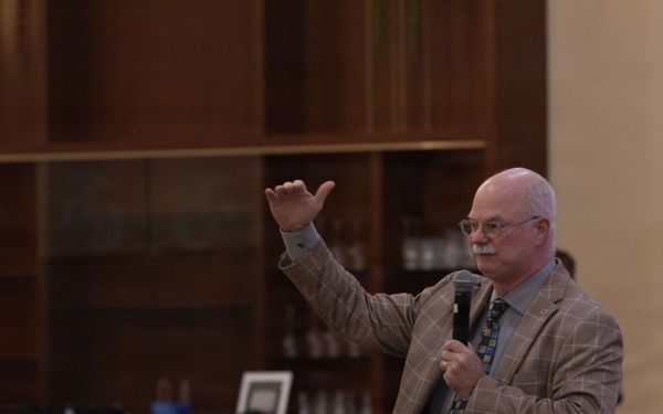 A middle-aged man in a plaid blazer and patterned tie is speaking to an audience while holding a microphone and gesturing with his raised right hand. The setting appears to be a formal dining hall or meeting room with wood-paneled shelving in the background. At least one person in military camouflage uniform is visible among the attendees.