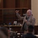A middle-aged man in a plaid blazer and patterned tie is speaking to an audience while holding a microphone and gesturing with his raised right hand. The setting appears to be a formal dining hall or meeting room with wood-paneled shelving in the background. At least one person in military camouflage uniform is visible among the attendees.