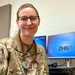 A female military medical personnel wearing U.S. Air Force OCP (Operational Camouflage Pattern) uniform with a stethoscope around her neck is posing for a photo in what appears to be a medical or administrative office. She is wearing glasses and has her hair pulled back. Behind her, a computer monitor displays the U.S. Department of Defense Defense Health Agency (DHA) login screen.