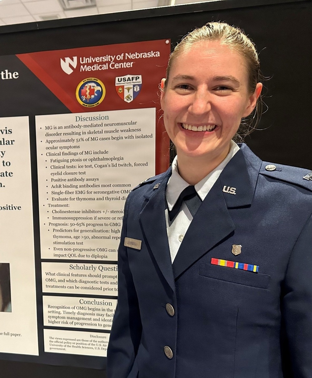 A person in a U.S. Air Force dress uniform is smiling in front of an academic research poster presentation. The poster is from the University of Nebraska Medical Center and discusses Myasthenia Gravis (MG), specifically Ocular Myasthenia Gravis (OMG). The individual appears to be presenting their scholarly research at a medical conference or symposium.