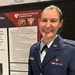 A person in a U.S. Air Force dress uniform is smiling in front of an academic research poster presentation. The poster is from the University of Nebraska Medical Center and discusses Myasthenia Gravis (MG), specifically Ocular Myasthenia Gravis (OMG). The individual appears to be presenting their scholarly research at a medical conference or symposium.