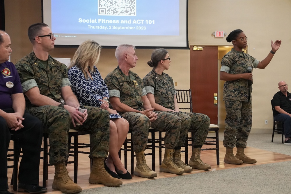 A military briefing or training session titled 'Social Fitness and ACT 101' is taking place on Thursday, 3 September 2026. A female Marine in camouflage uniform is standing and speaking into a microphone while gesturing, addressing a panel of seated individuals that includes both military personnel in MARPAT camouflage and at least one civilian woman. The event appears to be held in a formal meeting or conference hall.