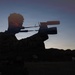A silhouetted individual wearing camouflage clothing is aiming a scoped rifle at dusk or dawn. The scene is set in an open, arid landscape with mountains visible in the background. A white pickup truck is parked in the background to the right.