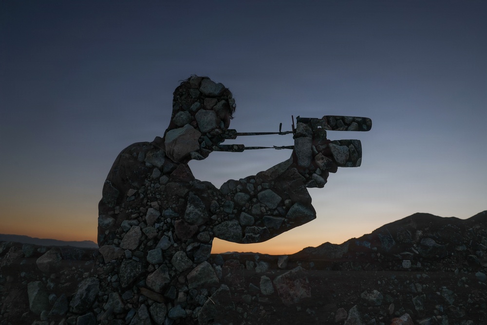A creative double-exposure or composite photograph blending the silhouette of a person operating a video camera with a rocky, stone-covered landscape. The image is set against a twilight sky with warm orange and cool blue tones near a mountainous horizon. The artistic technique makes the figure appear to be constructed entirely of rocks and stones.