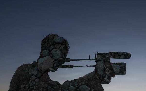 A creative double-exposure or composite photograph blending the silhouette of a person operating a video camera with a rocky, stone-covered landscape. The image is set against a twilight sky with warm orange and cool blue tones near a mountainous horizon. The artistic technique makes the figure appear to be constructed entirely of rocks and stones.