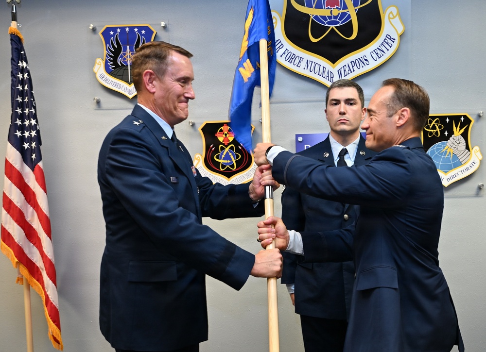 A military change of command ceremony is taking place at the Air Force Nuclear Weapons Center, where two officers in dress blue uniforms are exchanging a guidon (unit flag). A third uniformed airman stands at attention in the background. The setting is an official indoor military venue with unit insignia plaques mounted on the wall.