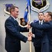 A military change of command ceremony is taking place at the Air Force Nuclear Weapons Center, where two officers in dress blue uniforms are exchanging a guidon (unit flag). A third uniformed airman stands at attention in the background. The setting is an official indoor military venue with unit insignia plaques mounted on the wall.
