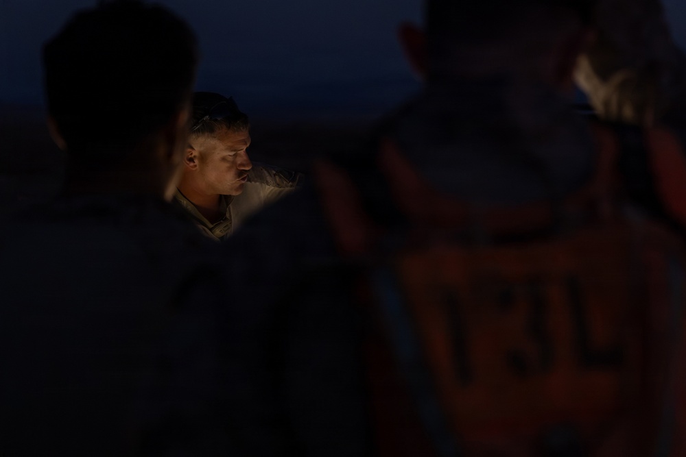 A nighttime scene showing a group of individuals gathered together, with one person's face illuminated by a light source in the center of the frame. The surrounding figures are largely silhouetted against a dark blue twilight sky. The image appears to capture a briefing or consultation occurring in low-light outdoor conditions.