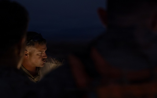 A nighttime scene showing a group of individuals gathered together, with one person's face illuminated by a light source in the center of the frame. The surrounding figures are largely silhouetted against a dark blue twilight sky. The image appears to capture a briefing or consultation occurring in low-light outdoor conditions.