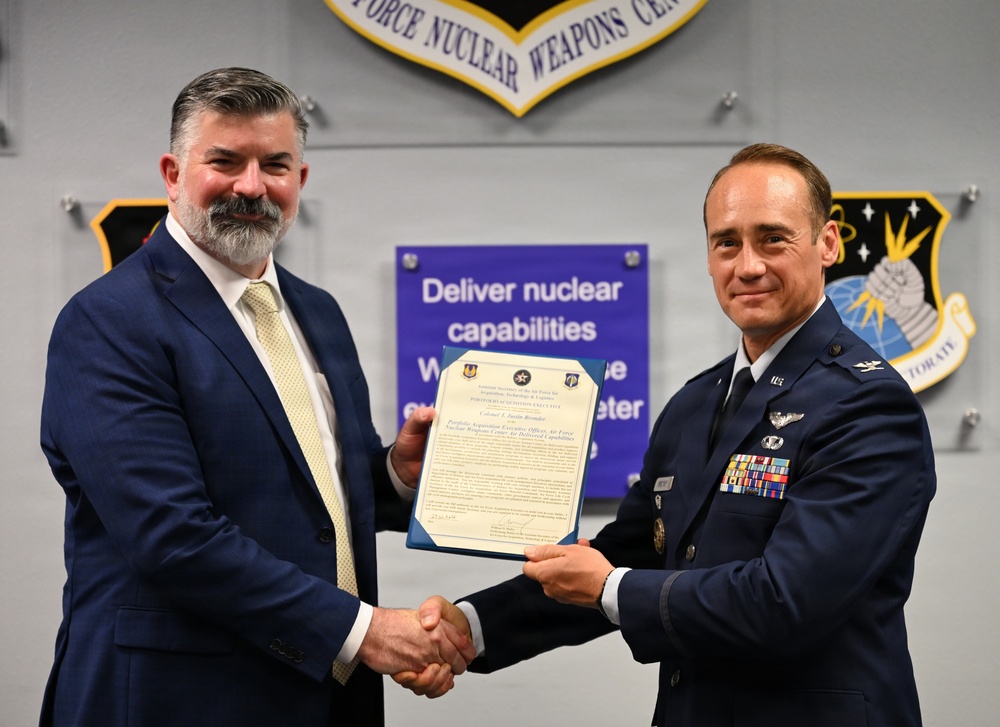 Two individuals are posing for a formal photograph at the Air Force Nuclear Weapons Center, shaking hands while jointly holding an official certificate or award document. One individual is in civilian attire (dark suit with yellow tie) and the other is in a U.S. Air Force dress blue uniform with the rank of Colonel and numerous service ribbons. The setting appears to be an official military recognition or designation ceremony.