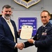 Two individuals are posing for a formal photograph at the Air Force Nuclear Weapons Center, shaking hands while jointly holding an official certificate or award document. One individual is in civilian attire (dark suit with yellow tie) and the other is in a U.S. Air Force dress blue uniform with the rank of Colonel and numerous service ribbons. The setting appears to be an official military recognition or designation ceremony.