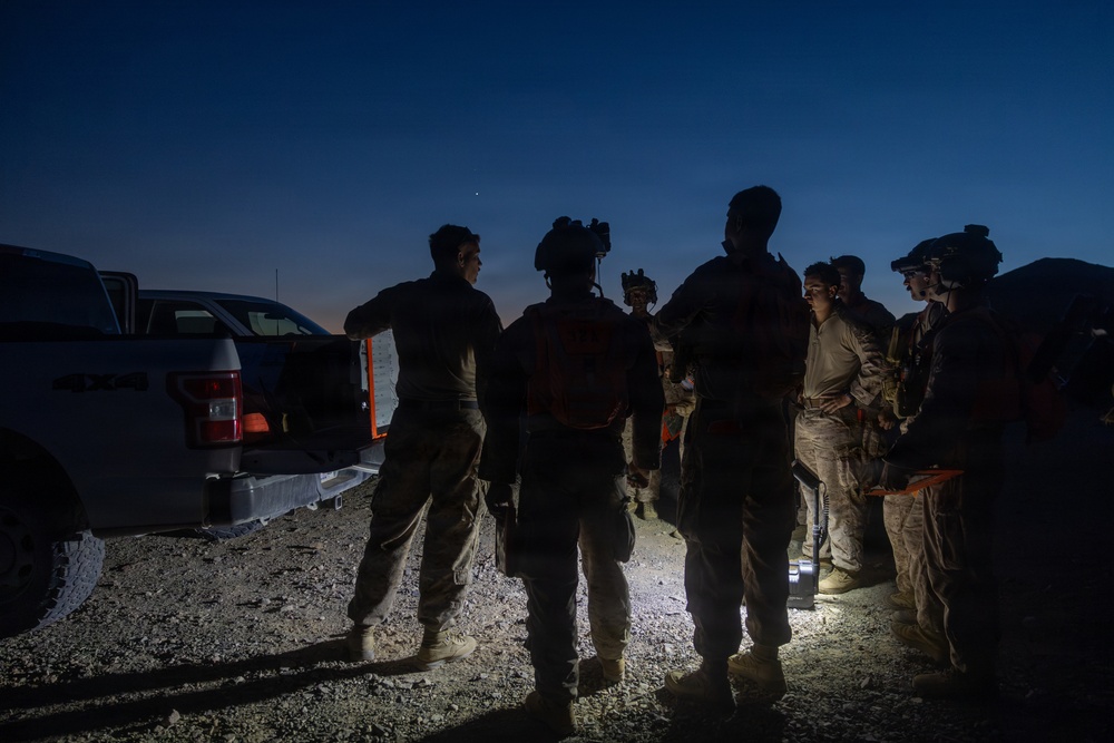 A group of military or tactical personnel are gathered outdoors at dusk or dawn near a pickup truck, appearing to conduct a briefing or pre-mission coordination. The scene is dimly lit, with a small ground-level light source illuminating the group from below. Personnel are equipped with tactical gear, helmets, and what appear to be night-vision devices.
