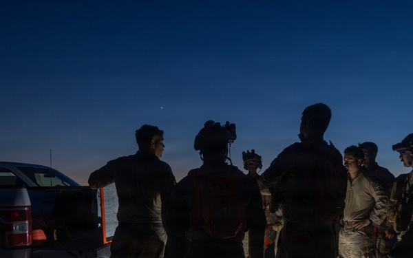 A group of military or tactical personnel are gathered outdoors at dusk or dawn near a pickup truck, appearing to conduct a briefing or pre-mission coordination. The scene is dimly lit, with a small ground-level light source illuminating the group from below. Personnel are equipped with tactical gear, helmets, and what appear to be night-vision devices.