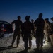 A group of military or tactical personnel are gathered outdoors at dusk or dawn near a pickup truck, appearing to conduct a briefing or pre-mission coordination. The scene is dimly lit, with a small ground-level light source illuminating the group from below. Personnel are equipped with tactical gear, helmets, and what appear to be night-vision devices.