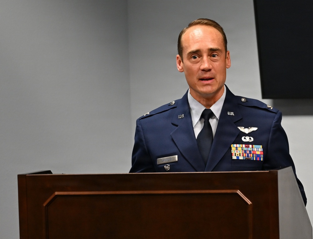 A U.S. Air Force officer in dress blues is speaking at a wooden podium. He displays multiple service ribbons, pilot wings, and a command badge on his uniform, indicating a senior officer rank. The setting appears to be a formal military ceremony or briefing room.