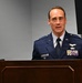 A U.S. Air Force officer in dress blues is speaking at a wooden podium. He displays multiple service ribbons, pilot wings, and a command badge on his uniform, indicating a senior officer rank. The setting appears to be a formal military ceremony or briefing room.