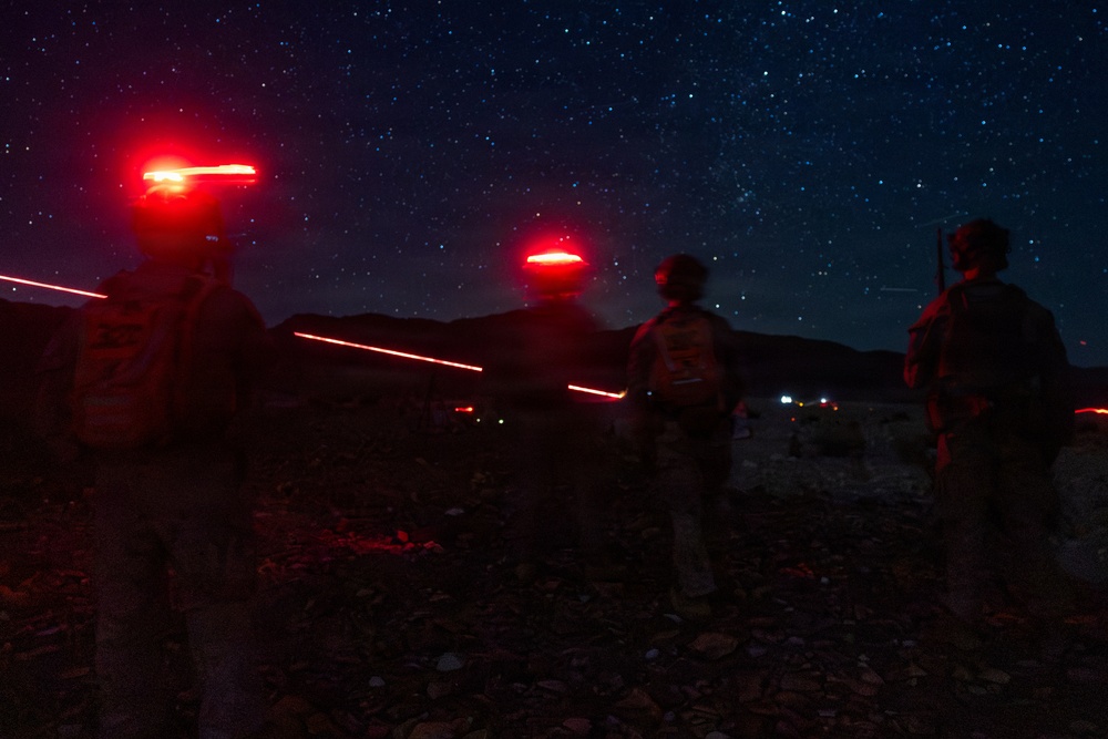 A nighttime outdoor scene showing multiple military personnel standing in a rocky, desert-like terrain under a clear, star-filled sky. The figures are equipped with tactical gear and are wearing red infrared/strobe marker lights on their helmets, which are glowing brightly. Additional lights are visible in the distant background, suggesting other personnel or vehicles in the area.