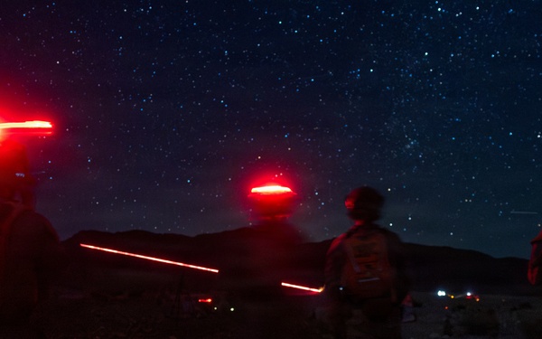 A nighttime outdoor scene showing multiple military personnel standing in a rocky, desert-like terrain under a clear, star-filled sky. The figures are equipped with tactical gear and are wearing red infrared/strobe marker lights on their helmets, which are glowing brightly. Additional lights are visible in the distant background, suggesting other personnel or vehicles in the area.
