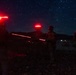 A nighttime outdoor scene showing multiple military personnel standing in a rocky, desert-like terrain under a clear, star-filled sky. The figures are equipped with tactical gear and are wearing red infrared/strobe marker lights on their helmets, which are glowing brightly. Additional lights are visible in the distant background, suggesting other personnel or vehicles in the area.