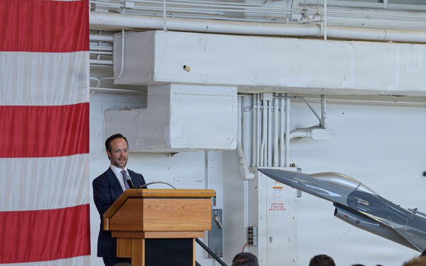 A civilian male in a dark suit is delivering a speech at a wooden podium inside a military hangar. The audience consists primarily of uniformed military personnel in camouflage uniforms, along with some civilians. A large American flag is displayed on the left, and an F-16 fighter jet is visible in the background.