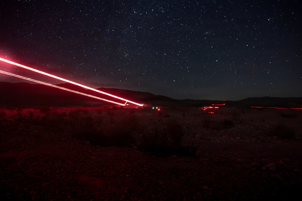 A long-exposure night photograph taken in a desert landscape showing vehicle light trails streaking diagonally across the frame against a star-filled sky. Red tail lights from passing vehicles have created bright, continuous streaks due to the extended shutter speed. The scene captures a remote desert road with mountains silhouetted on the horizon.