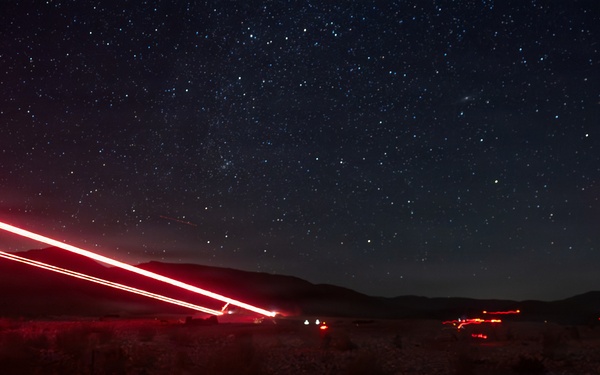 A long-exposure night photograph taken in a desert landscape showing vehicle light trails streaking diagonally across the frame against a star-filled sky. Red tail lights from passing vehicles have created bright, continuous streaks due to the extended shutter speed. The scene captures a remote desert road with mountains silhouetted on the horizon.