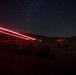 A long-exposure night photograph taken in a desert landscape showing vehicle light trails streaking diagonally across the frame against a star-filled sky. Red tail lights from passing vehicles have created bright, continuous streaks due to the extended shutter speed. The scene captures a remote desert road with mountains silhouetted on the horizon.