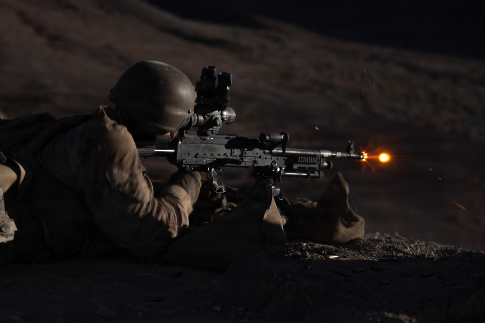 A soldier in prone firing position is discharging a light machine gun at night, with a visible muzzle flash illuminating the scene. The shooter is equipped with tactical gear including a helmet with mounted optics and is using a bipod-supported weapon. The image was captured in low-light conditions with the muzzle flash serving as the primary light source.