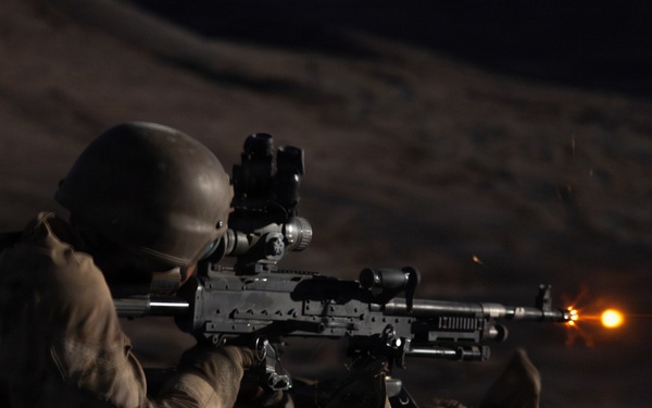 A soldier in prone firing position is discharging a light machine gun at night, with a visible muzzle flash illuminating the scene. The shooter is equipped with tactical gear including a helmet with mounted optics and is using a bipod-supported weapon. The image was captured in low-light conditions with the muzzle flash serving as the primary light source.