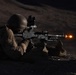 A soldier in prone firing position is discharging a light machine gun at night, with a visible muzzle flash illuminating the scene. The shooter is equipped with tactical gear including a helmet with mounted optics and is using a bipod-supported weapon. The image was captured in low-light conditions with the muzzle flash serving as the primary light source.