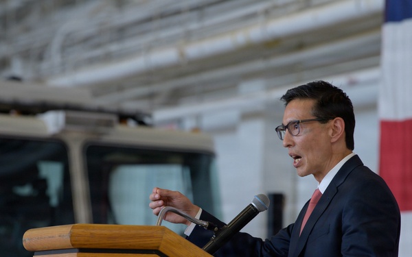 A man in a dark suit with a pink/salmon tie is speaking at a wooden podium equipped with a microphone. The event appears to be an official or civic ceremony held inside a fire station, as evidenced by a fire truck visible in the background. An American flag is displayed prominently to the right of the speaker.