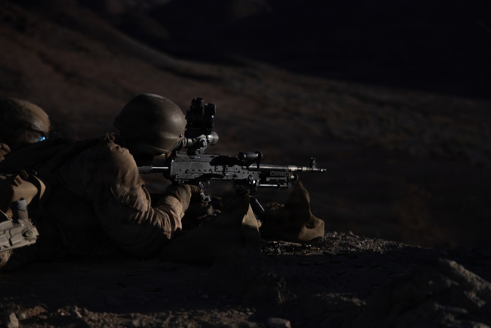 A soldier in full combat gear is photographed in a prone firing position at night, aiming a light machine gun equipped with an optical scope. The image appears to be taken during a nighttime military training exercise or combat operation in an arid, rocky terrain. A second soldier is partially visible on the left side of the frame.