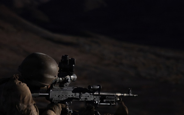 A soldier in full combat gear is photographed in a prone firing position at night, aiming a light machine gun equipped with an optical scope. The image appears to be taken during a nighttime military training exercise or combat operation in an arid, rocky terrain. A second soldier is partially visible on the left side of the frame.