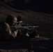 A soldier in full combat gear is photographed in a prone firing position at night, aiming a light machine gun equipped with an optical scope. The image appears to be taken during a nighttime military training exercise or combat operation in an arid, rocky terrain. A second soldier is partially visible on the left side of the frame.