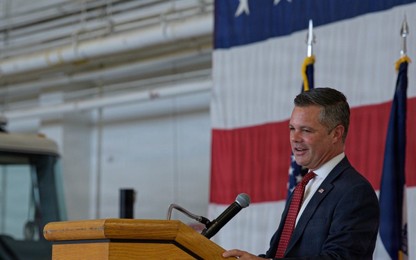 A man in a dark suit with a red tie is speaking at a wooden podium equipped with a microphone. The setting appears to be a fire station bay, evidenced by a fire truck visible in the background. An American flag and what appears to be a state flag are displayed behind the speaker.