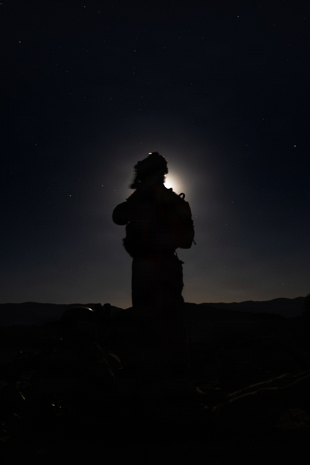A nighttime silhouette photograph showing a single person standing against a moonlit sky with visible stars. The subject appears to be wearing cold-weather gear including a fur-trimmed hat and carrying a backpack, positioned on elevated terrain. A bright light source — likely the moon — creates a halo effect directly behind the subject's head.