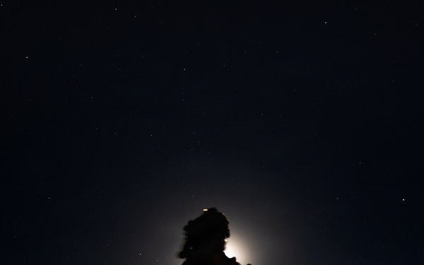 A nighttime silhouette photograph showing a single person standing against a moonlit sky with visible stars. The subject appears to be wearing cold-weather gear including a fur-trimmed hat and carrying a backpack, positioned on elevated terrain. A bright light source — likely the moon — creates a halo effect directly behind the subject's head.