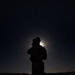 A nighttime silhouette photograph showing a single person standing against a moonlit sky with visible stars. The subject appears to be wearing cold-weather gear including a fur-trimmed hat and carrying a backpack, positioned on elevated terrain. A bright light source — likely the moon — creates a halo effect directly behind the subject's head.