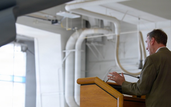 A man in an olive/tan suit is delivering a speech at a wooden podium inside what appears to be a military aircraft hangar. The audience consists primarily of uniformed military personnel seated in rows, along with a few civilians in business attire. The setting suggests a formal military ceremony or official address.