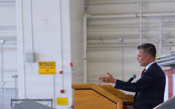 A civilian official in a dark suit is delivering a speech at a wooden podium inside a military aircraft hangar. He is addressing a seated audience of military personnel in camouflage uniforms. A fighter jet with markings '132 FW' and 'IA' (Iowa Air National Guard, 132nd Fighter Wing) is prominently visible in the foreground.