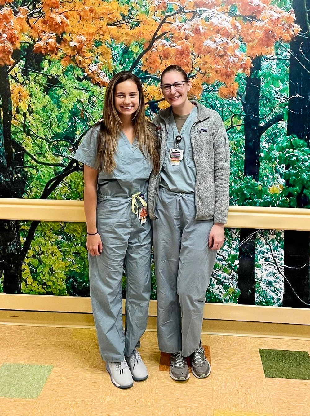 Two individuals wearing matching gray/teal scrubs are posing together and smiling in what appears to be a hospital or medical facility hallway. Both are wearing ID badges, and one has a stethoscope and is wearing a gray Patagonia fleece jacket over her scrubs. They are standing in front of a large decorative mural depicting an autumn forest scene.