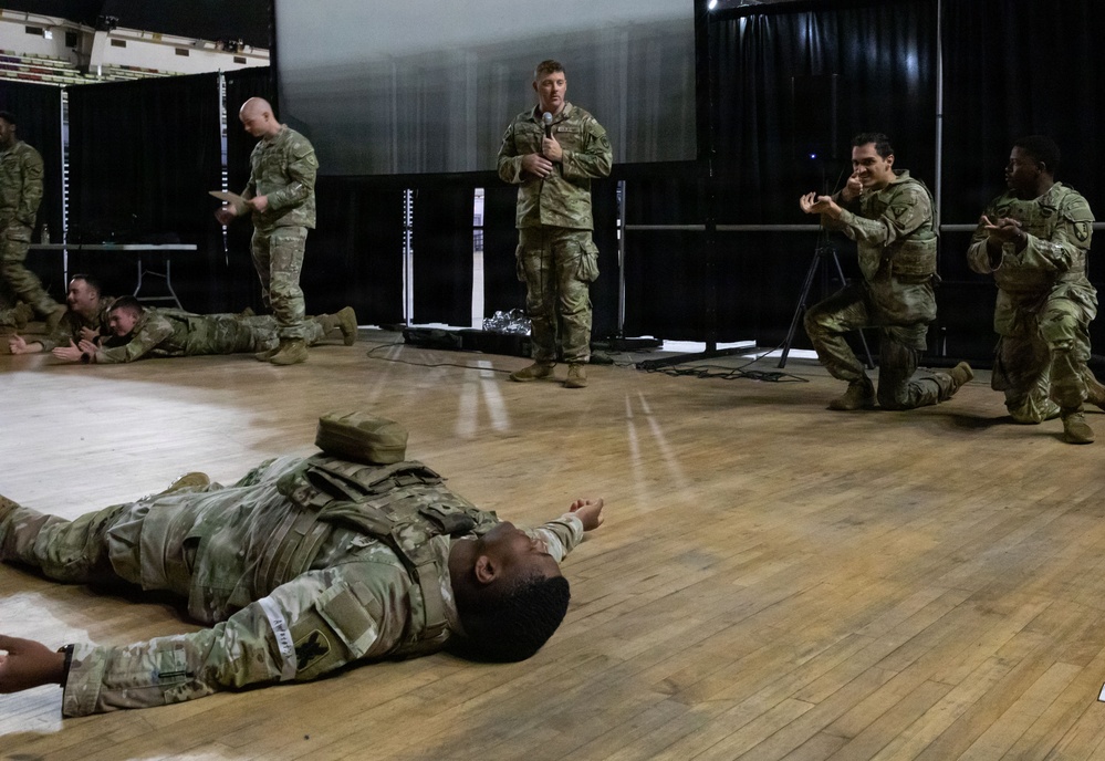 A group of military personnel in OCP (Operational Camouflage Pattern) uniforms are engaged in what appears to be a training exercise or demonstration inside an indoor facility with a wooden floor and stage area. One soldier stands centrally holding a microphone, apparently instructing or narrating, while other soldiers are positioned on the floor or in crouching stances around him. The scene suggests a combat casualty care, tactical movement, or scenario-based training demonstration.