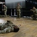 A group of military personnel in OCP (Operational Camouflage Pattern) uniforms are engaged in what appears to be a training exercise or demonstration inside an indoor facility with a wooden floor and stage area. One soldier stands centrally holding a microphone, apparently instructing or narrating, while other soldiers are positioned on the floor or in crouching stances around him. The scene suggests a combat casualty care, tactical movement, or scenario-based training demonstration.