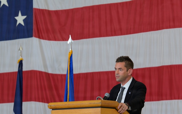 A man in a dark suit and tie is speaking at a wooden podium during what appears to be an official military or government ceremony. A large American flag serves as a backdrop, flanked by additional flags including what appears to be a U.S. Air Force flag. Several audience members, some in military uniforms, are visible in the foreground with their backs to the camera.