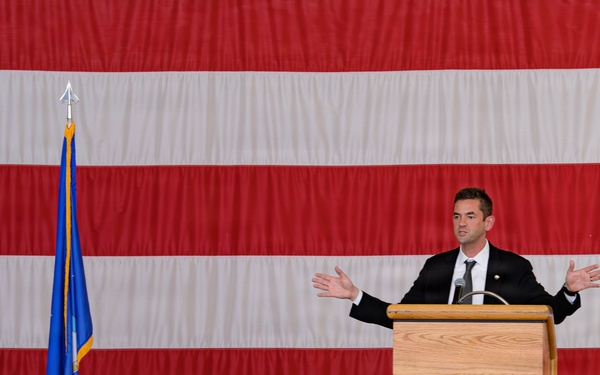 A civilian male in a dark suit is delivering a speech at a wooden podium before a large audience of military personnel in camouflage uniforms. The setting appears to be a military hangar or large indoor facility, decorated with an oversized American flag as a backdrop. Two flags on poles — one appearing to be a state flag and one a blue military branch flag — flank the speaker.